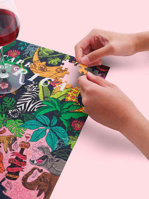 Nahaufnahme eines Wein-Puzzles mit Südafrika-Motiven, das von zwei Händen zusammengesetzt wird. Neben dem Puzzle steht ein Glas Rotwein.