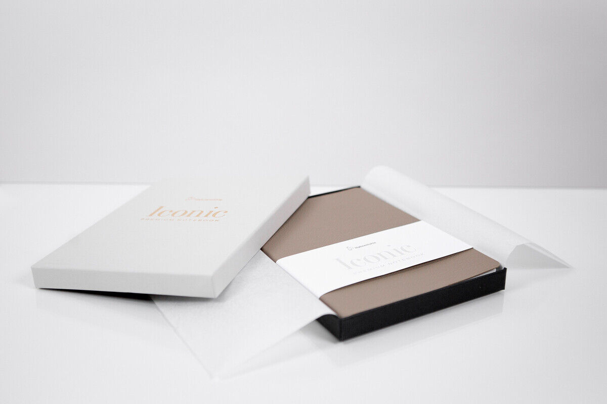 Ein elegantes Notizbuch im DIN A5 Format, verpackt in einer weißen Box mit der Aufschrift 'iconic'. Das Notizbuch hat einen beigen Einband und eine weiße Banderole, die das Buch zusammenhält. Die Szene ist minimalistisch und modern gestaltet.