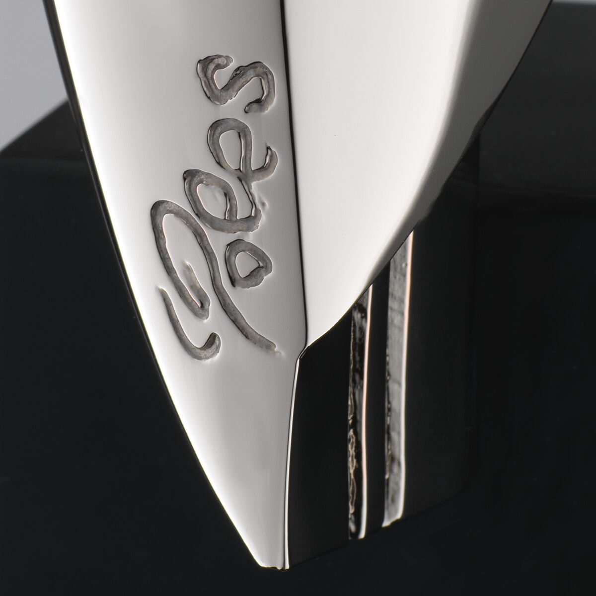 Detailaufnahme einer silberfarbenen, glänzenden Skulptur aus Kunstguss mit stilisierter Gravur des Namens 'Dobes'. Das Werk zeigt klare, glatte Oberflächen sowie eine moderne, elegante Form. Im Hintergrund dunkle, matte Fläche. Die Skulptur wirkt edel und hochwertig.