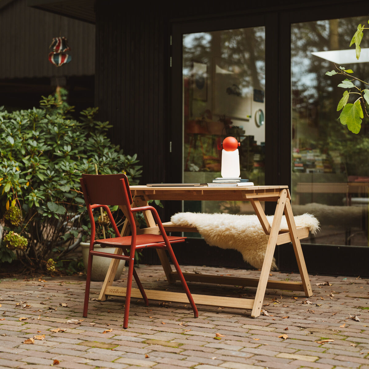 Ein klappbarer Tischbank-Set namens 'Folding Picnic Table' der Marke Weltevree steht auf einer Terrasse aus Pflastersteinen vor einem modernen Haus mit großen Glasfronten. Auf dem Tisch steht eine weiße Vase mit rotem Deckel. Ein roter Stuhl und eine Bank mit Schaffellauflage am Tisch. Viel grünes Laub umgibt die Szene.