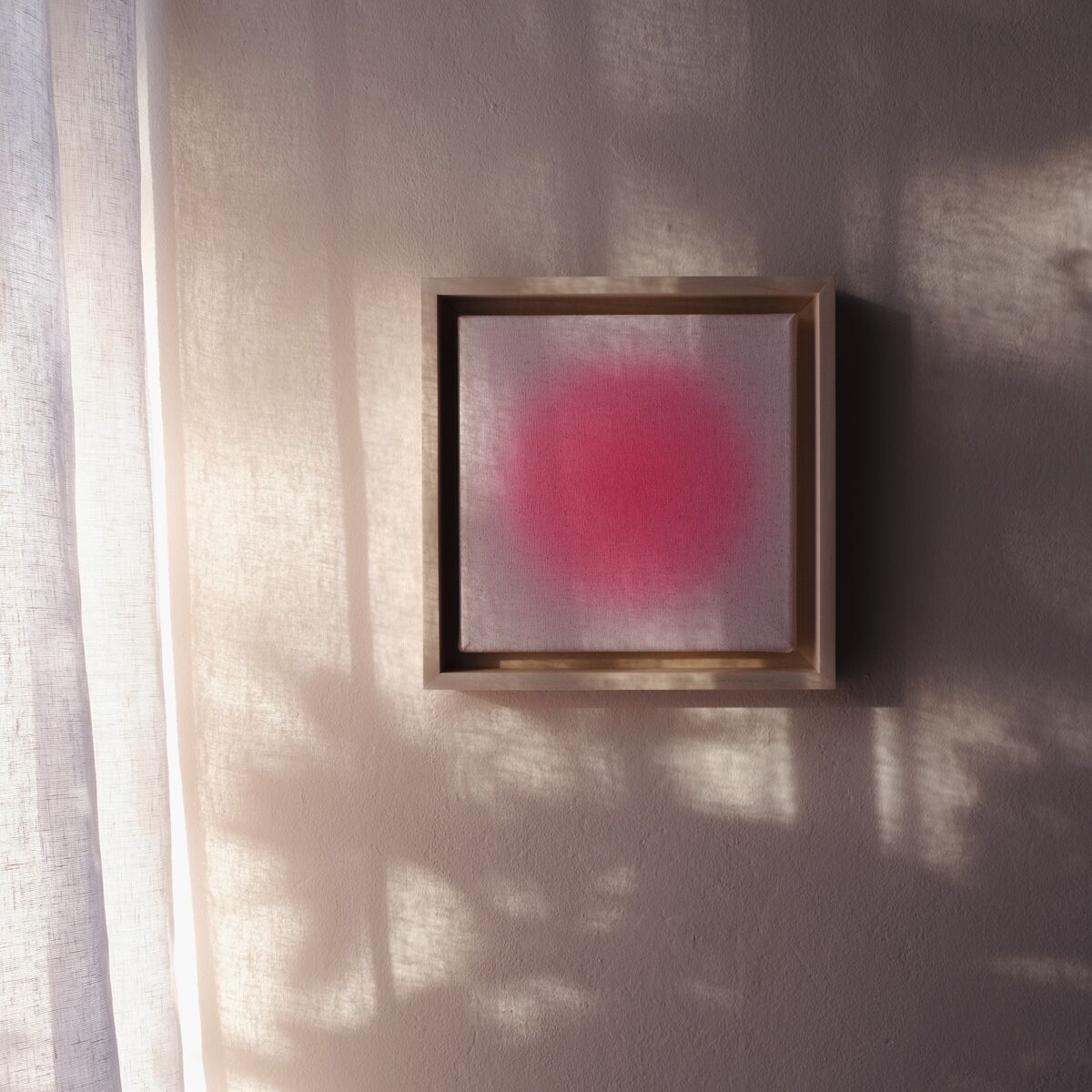 Quadratisches, minimalistisch gehaltenes Kunstwerk mit einem pinken Farbverlauf in der Mitte und hellem Hintergrund, eingerahmt in einem schlichten Holzrahmen. Das Bild hängt an einer hellen Wand, durch die Tageslicht und sanfte Schatten fallen.