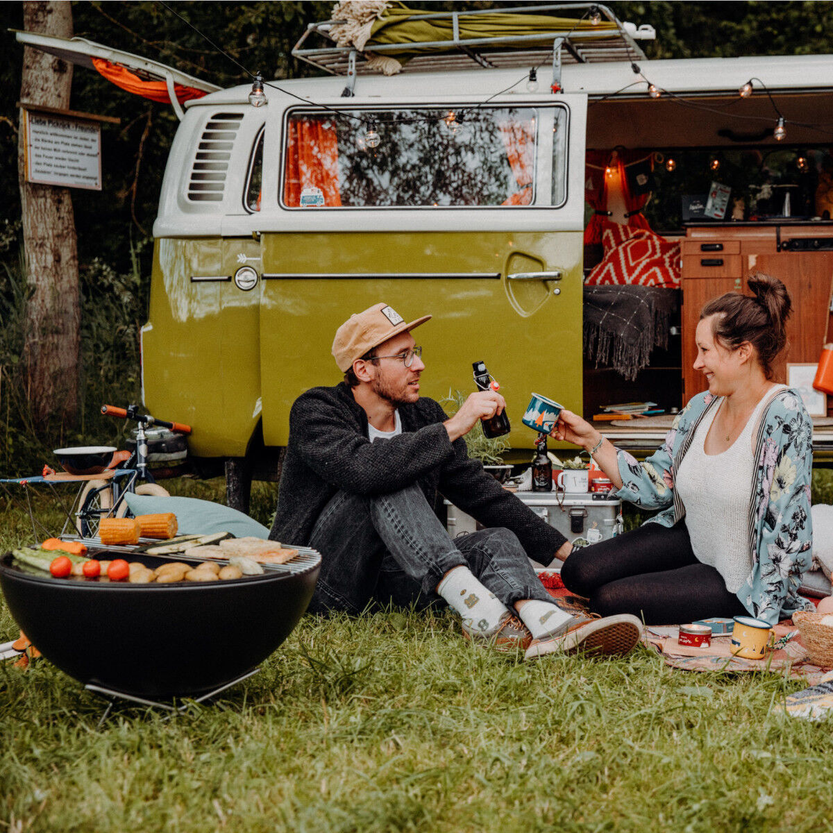 Zwei Personen sitzen auf einer Wiese neben einem Van, während sie um einen »BOWL« Plancha Halbring herum grillen.