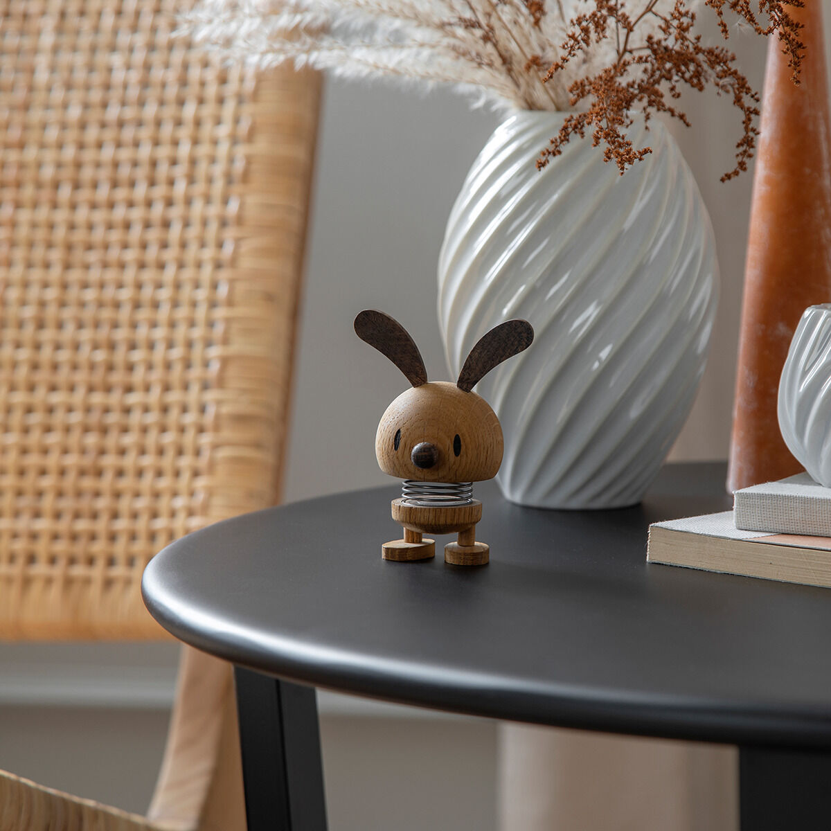 Eine dekorative Holzfigur in Form eines Hasen mit runden Ohren steht auf einem Tisch. Die Hoptimist »Bunny« Eiche Figur hat einen minimalistischen, skandinavischen Stil. Im Hintergrund sind ein dekorativer weißer Keramikvase und ein geflochtener Stuhl sichtbar.