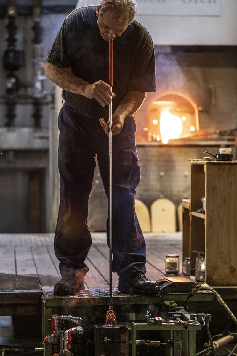 Ein Glasbläser arbeitet in einer Werkstatt, umgeben von verschiedenen Werkzeugen und Geräten. Im Hintergrund ist ein brennender Ofen zu sehen.