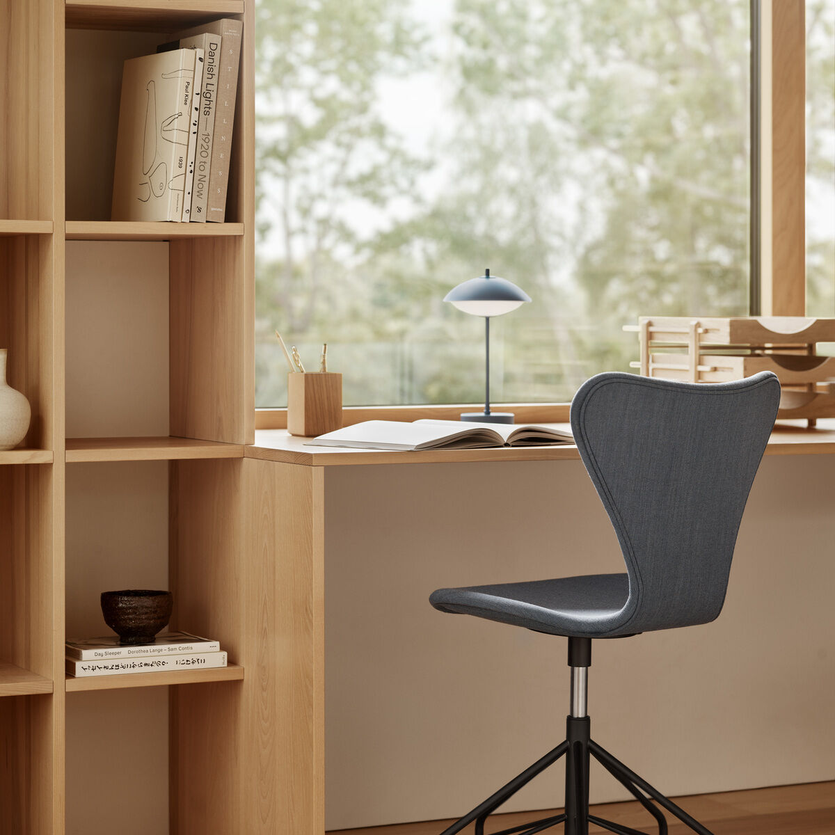 Modern eingerichteter Arbeitsplatz mit hellem Holzschreibtisch und -regal, darauf eine tragbare blaue Lampe »Clam™« von Fritz Hansen, Bücher, Stiftebox und Accessoires. Ein drehbarer, gepolsterter Bürostuhl auf Rollen steht am Tisch. Große Fenster ermöglichen einen Blick ins Grüne.