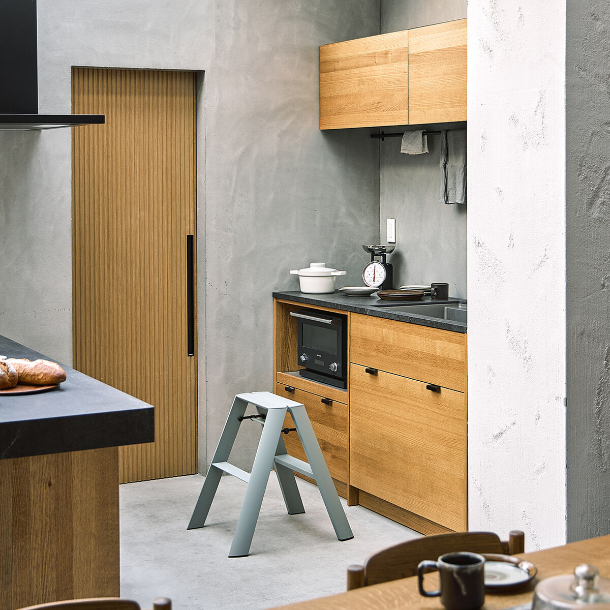 Eine moderne zweistufige Lucano Trittleiter steht in einer hellen, minimalistischen Küche mit Holzschränken und grauen Wänden. Die Trittleiter ist hellgrau und platzsparend vor einer Arbeitsplatte positioniert, um das Erreichen höherer Regale zu erleichtern.