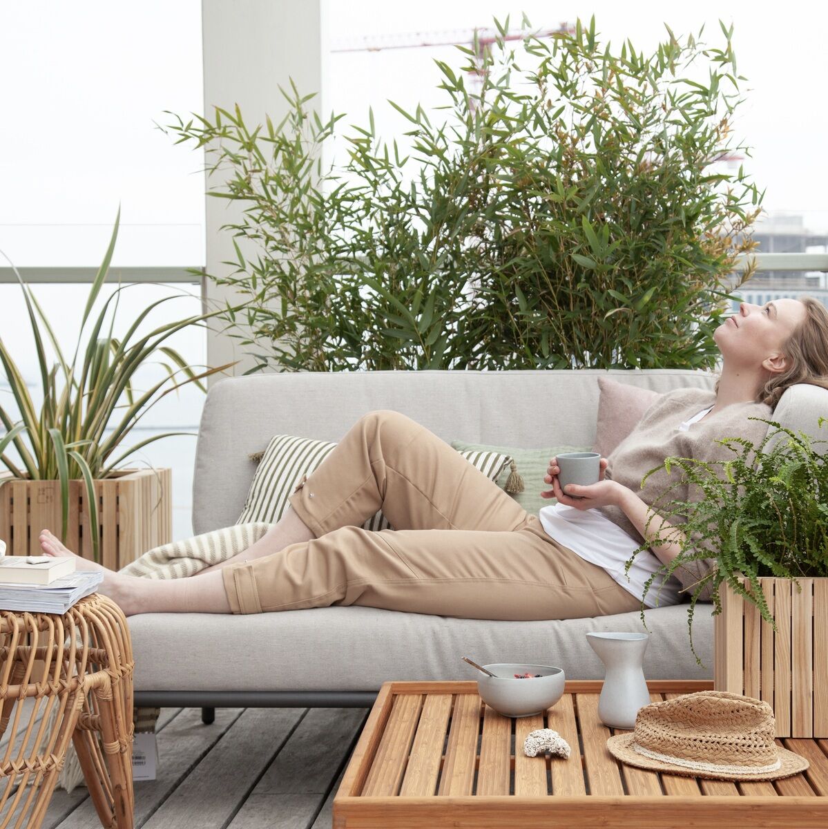 Eine Person liegt entspannt auf einem hellgrauen Outdoor-Sofa, umgeben von mehreren Pflanzgefäßen »GrowMORE« aus Holz mit großen grünen Pflanzen. Ein Rattan-Tisch und Holzelemente ergänzen die gemütliche Atmosphäre auf dem Balkon oder der Terrasse.