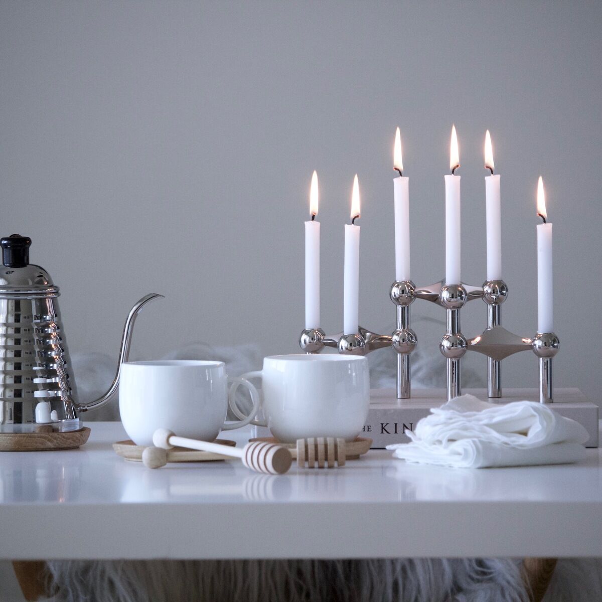 Ein moderner Kerzenständer aus Chrom mit sieben weißen Kerzen auf einem Esstisch, gestaltet mit weißen Tassen, einer Teekanne und Tischdekoration, in einem minimalistisch eingerichteten Raum.