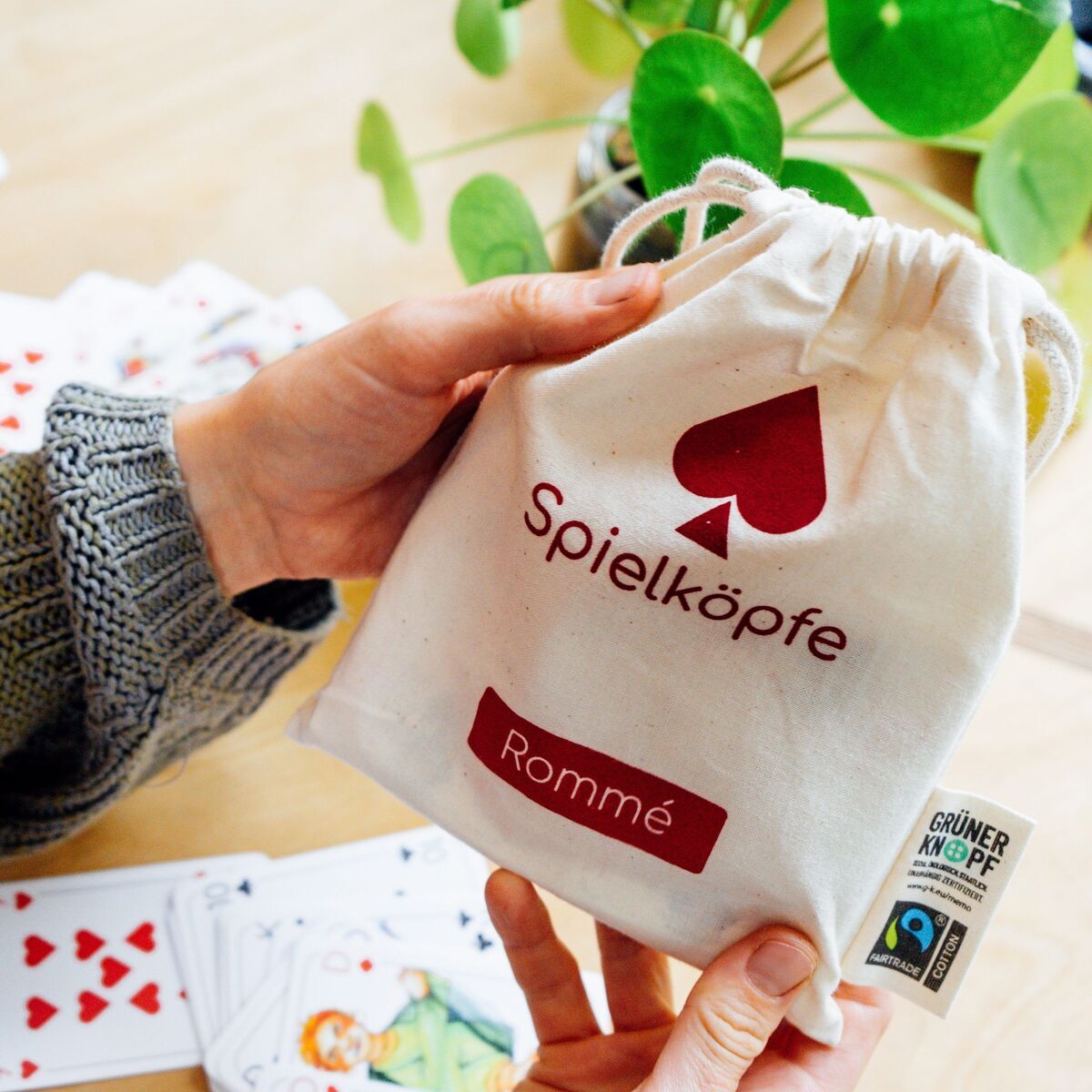 Eine Textiltasche mit der Aufschrift 'Spielköpfe Rommé' wird von einer Person gehalten. Im Hintergrund liegen Spielkarten auf einem Tisch, und im Bild sind auch grüne Blätter einer Pflanze zu sehen.