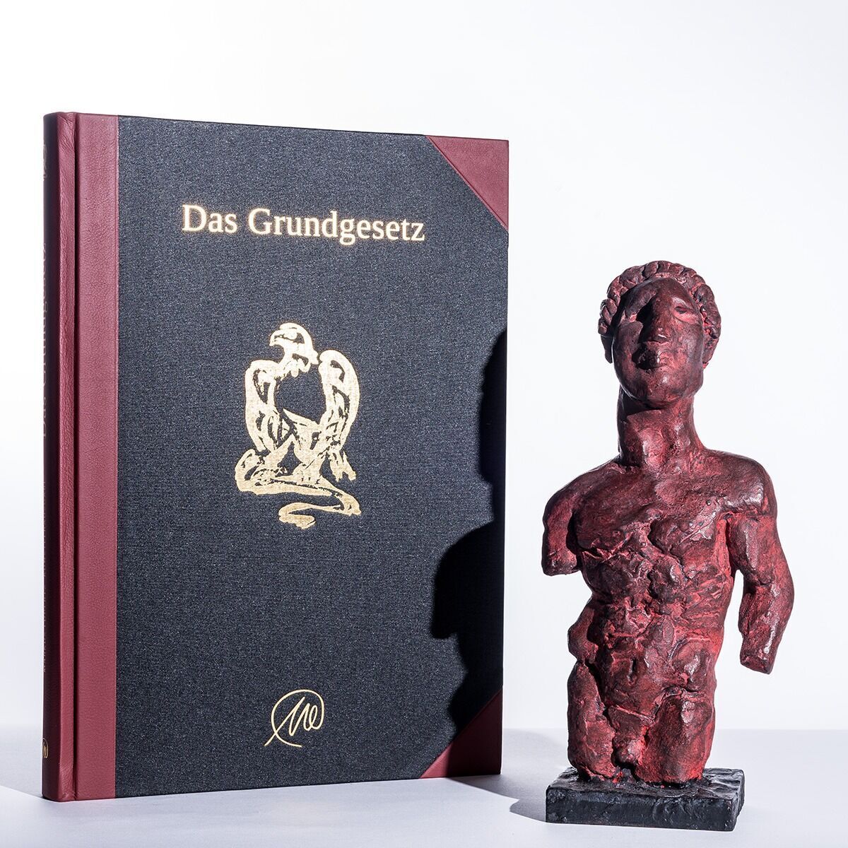 Ein Kunstbuch mit dem Titel 'Das Grundgesetz' von Markus Lüpertz aus dem Jahr 2013 steht neben einer rot bemalten Skulptur. Das Buch hat eine schwarze Frontseite mit einem goldenen Adler und eine rote Umrandung. Die Skulptur zeigt einen menschlichen Oberkörper in einer abstrakten Form.