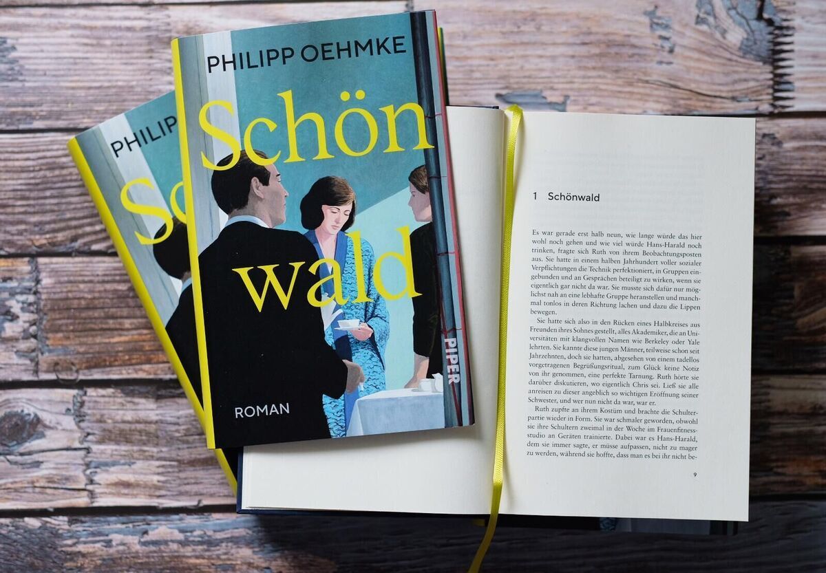 Zwei Exemplare des Buches 'Schönwald' von Philipp Oehmke liegen geschlossen auf einem Holztisch, daneben ein geöffnetes Exemplar mit gut lesbaren Buchseiten und einem gelben Lesezeichenband.