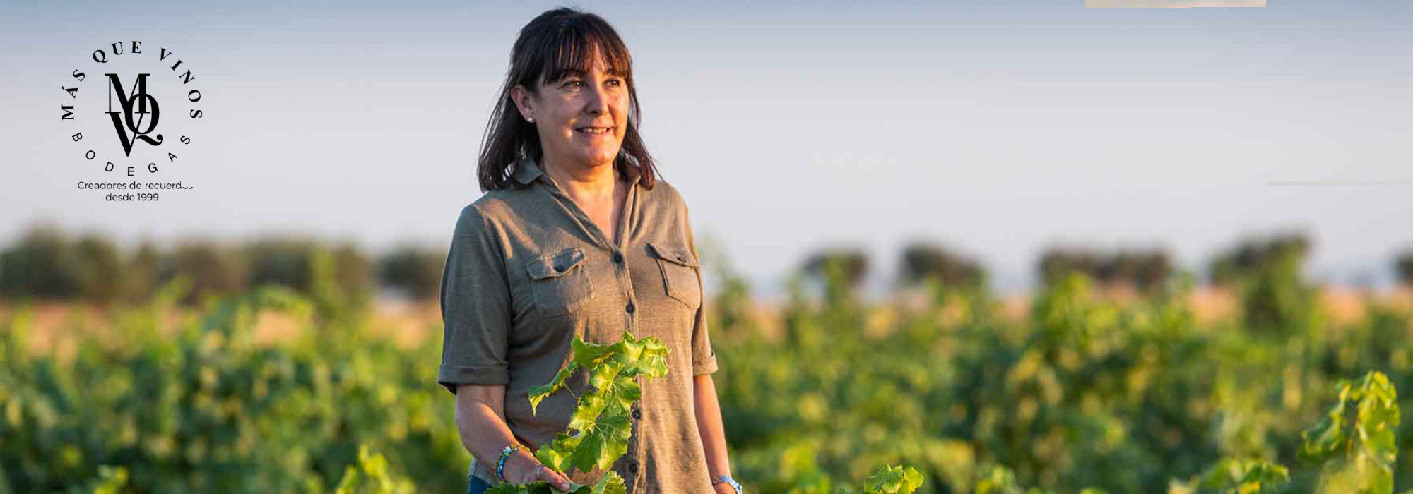 Frau in khakifarbenem Hemd steht in einem Weinberg und hält eine Rebe in der Hand, Logo von Mas Que Vinos links oben