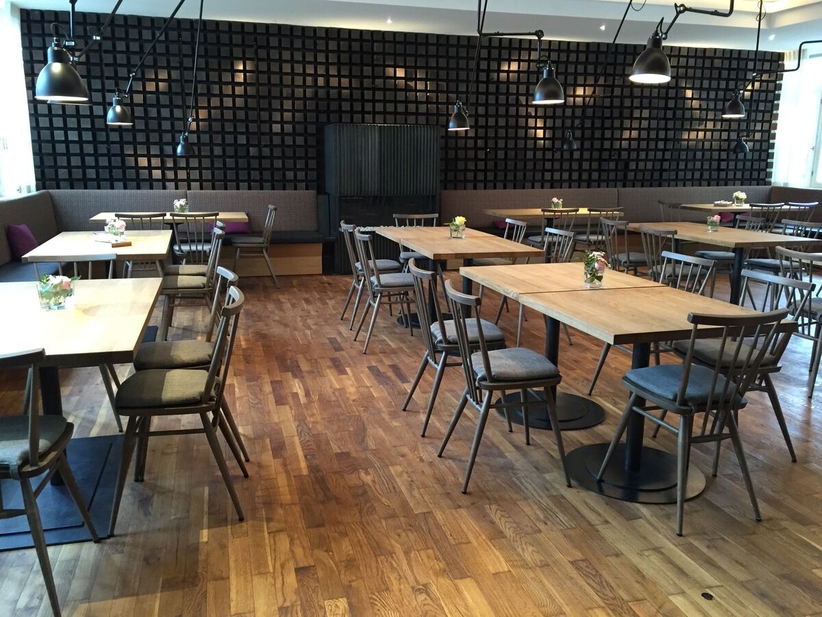 Ein modernes Restaurant mit Holzboden und Holztischen, die von Ercol-Stühlen in grau gebeizt umgeben sind. Die Stühle sind schlicht gestaltet und passen gut zum skandinavischen Design des Raumes. Mehrere Blumenarrangements stehen auf den Tischen als Dekoration.