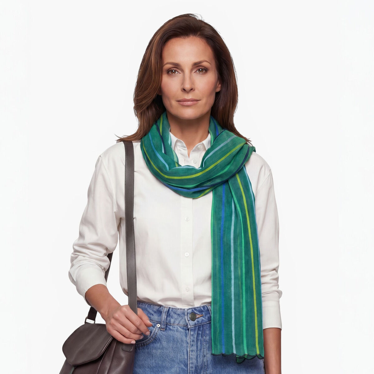 Eine Frau trägt einen gestreiften Leinenschal in Grün und bunten Tönen von Ulrike Isensee, kombiniert mit einer weißen Bluse, blauer Jeans und einer braunen Umhängetasche. Der Hintergrund ist neutral weiß. Der Schal wirkt leicht und modisch.
