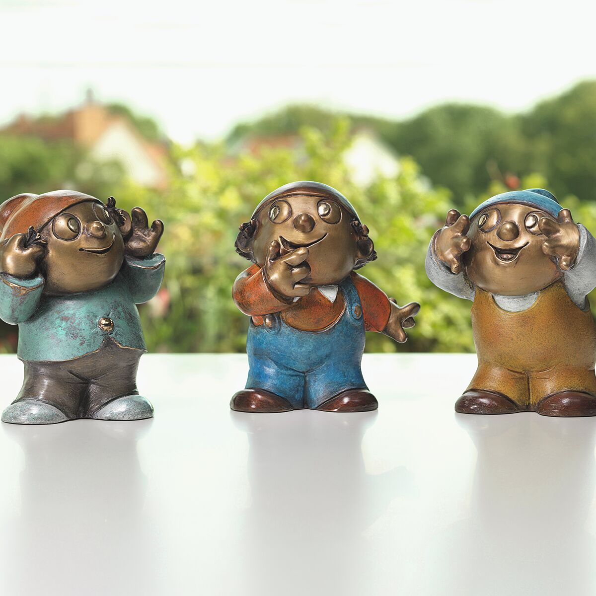 Drei detailreiche, farbig patinierte Bronzefiguren der bekannten Mainzelmännchen stehen nebeneinander auf einer glatten, hellen Fläche vor grün verschwommener Naturkulisse. Die Figuren tragen verschiedene bunte Outfits und zeigen unterschiedliche, freundliche Gesten.