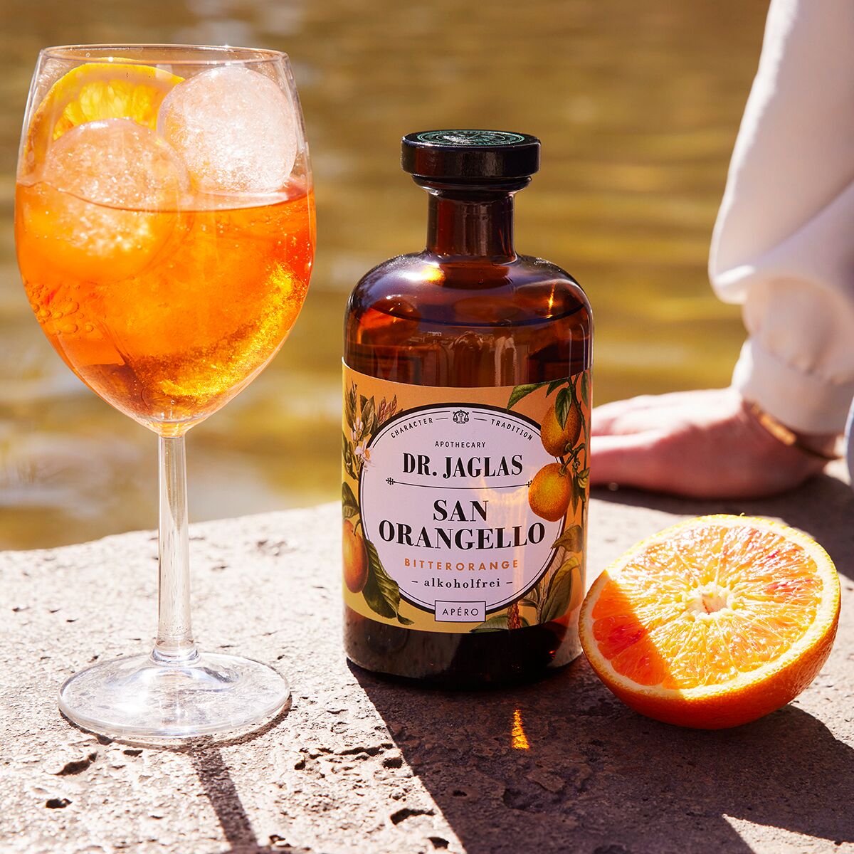 Eine stilvolle Szene am Wasser mit einem Glas orangefarbenem, eisgekühltem Aperitif, einer Flasche "San Orangello alkoholfreier Bitterorangen-Aperitif" von Dr. Jaglas und einer aufgeschnittenen Orange auf hellem Stein. Im Hintergrund liegt ein Arm entspannt auf dem Boden.