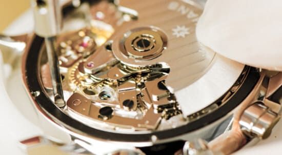 Nahaufnahme des komplizierten mechanischen Uhrwerks mit seinen goldenen Zahnrädern und Edelsteinen, das ein Techniker präzise einstellt.