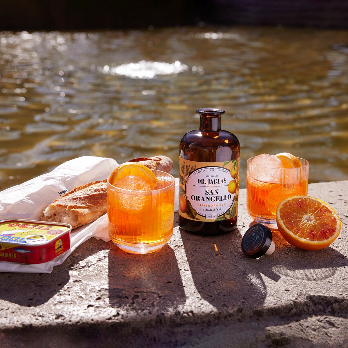 Auf einer Steinfläche am Wasser stehen eine braune Flasche San Orangello alkoholfreier Bitterorangen-Aperitif von Dr. Jaglas, zwei Gläser mit orangefarbenem Getränk und Eis, eine aufgeschnittene Orange, Baguette und eine offene Sardinendose, alles sonnendurchflutet und einladend arrangiert.