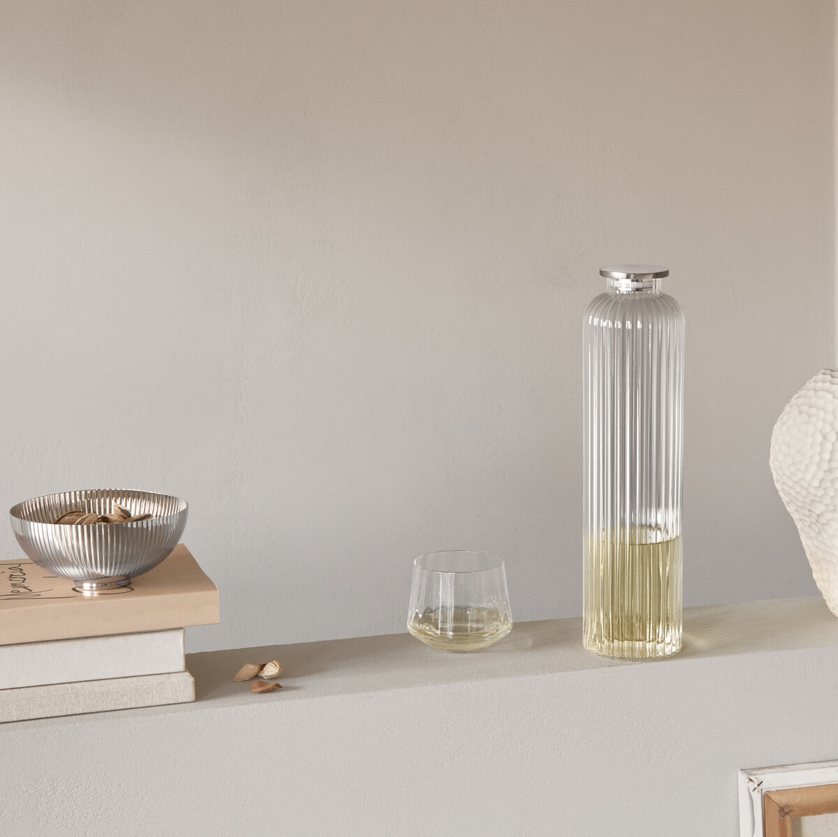 Das Bild zeigt eine moderne und elegante Karaffe »Bernadotte« zusammen mit einem Glas und verschiedenen Dekorationselementen auf einem Regal. Die Farbgebung ist hauptsächlich in Silber, Transparent, Weiß und Hellbeige gehalten.