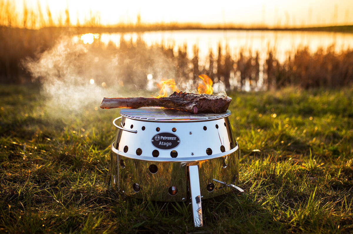 Der Grill »Atago« steht auf einer grünen Wiese am See bei Sonnenuntergang. Auf dem Grillrost brennt ein Steak, und Rauch steigt auf.