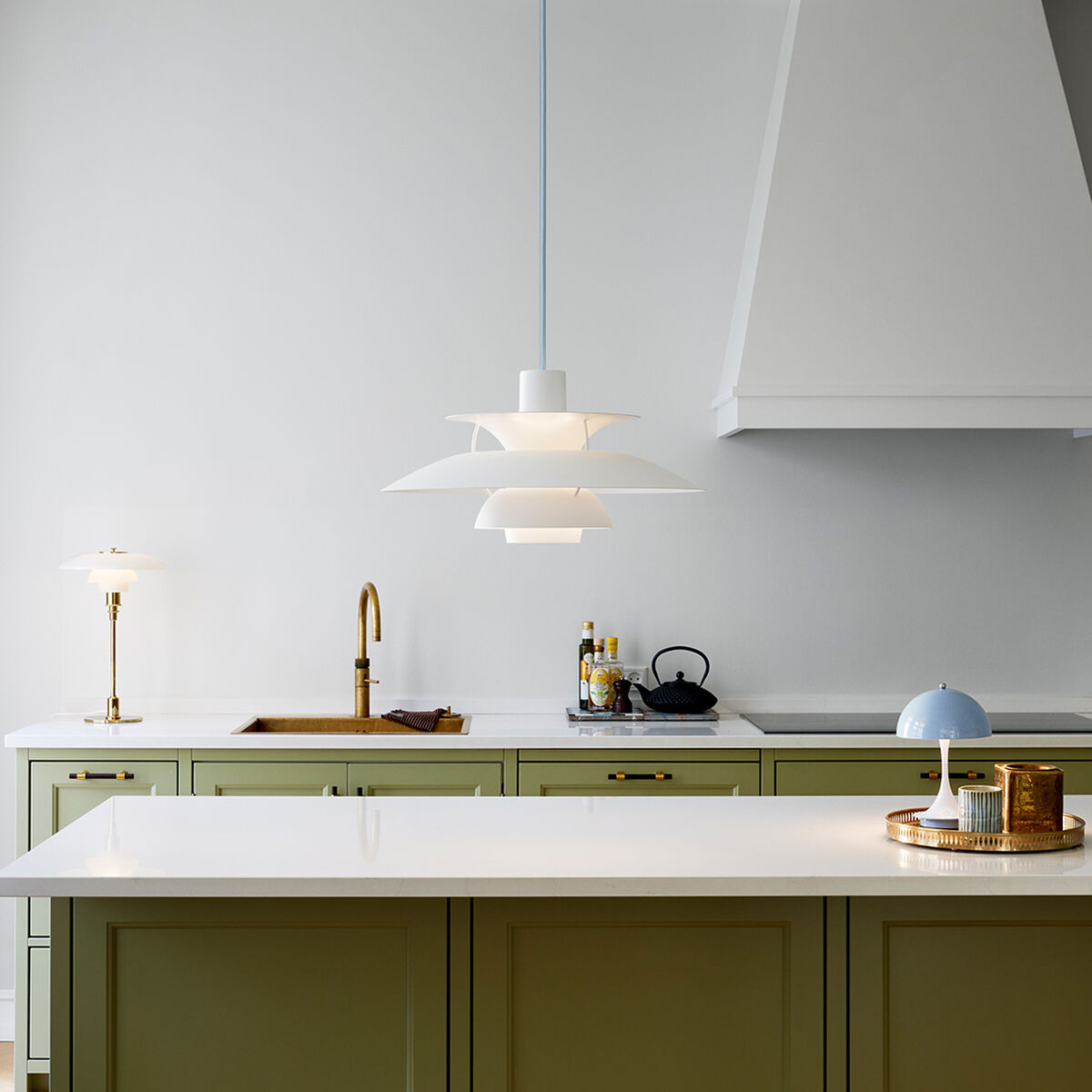 Moderne Küche mit salbeigrüner Kücheninsel und heller Arbeitsplatte. Im Zentrum hängt die ZEIT-Sonderedition Pendelleuchte »PH5 Monochrome« Blaugrau. Die Szene ist harmonisch und minimalistisch gestaltet mit goldenen Armaturen und dezenten Accessoires auf hellem Parkett.