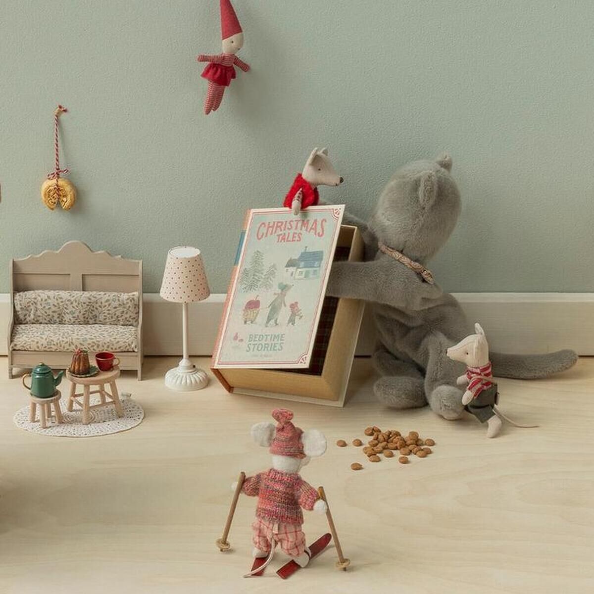 Mehrere Maileg-Stofftiere, darunter ein großes Schwein und verschiedene weihnachtlich gekleidete Mäuse, sitzen auf hellem Boden vor einer mintgrünen Wand. Sie sind um Miniaturmöbel, ein Buch und kleine Dekorationen arrangiert und bilden eine festliche, winterliche Spielszene.