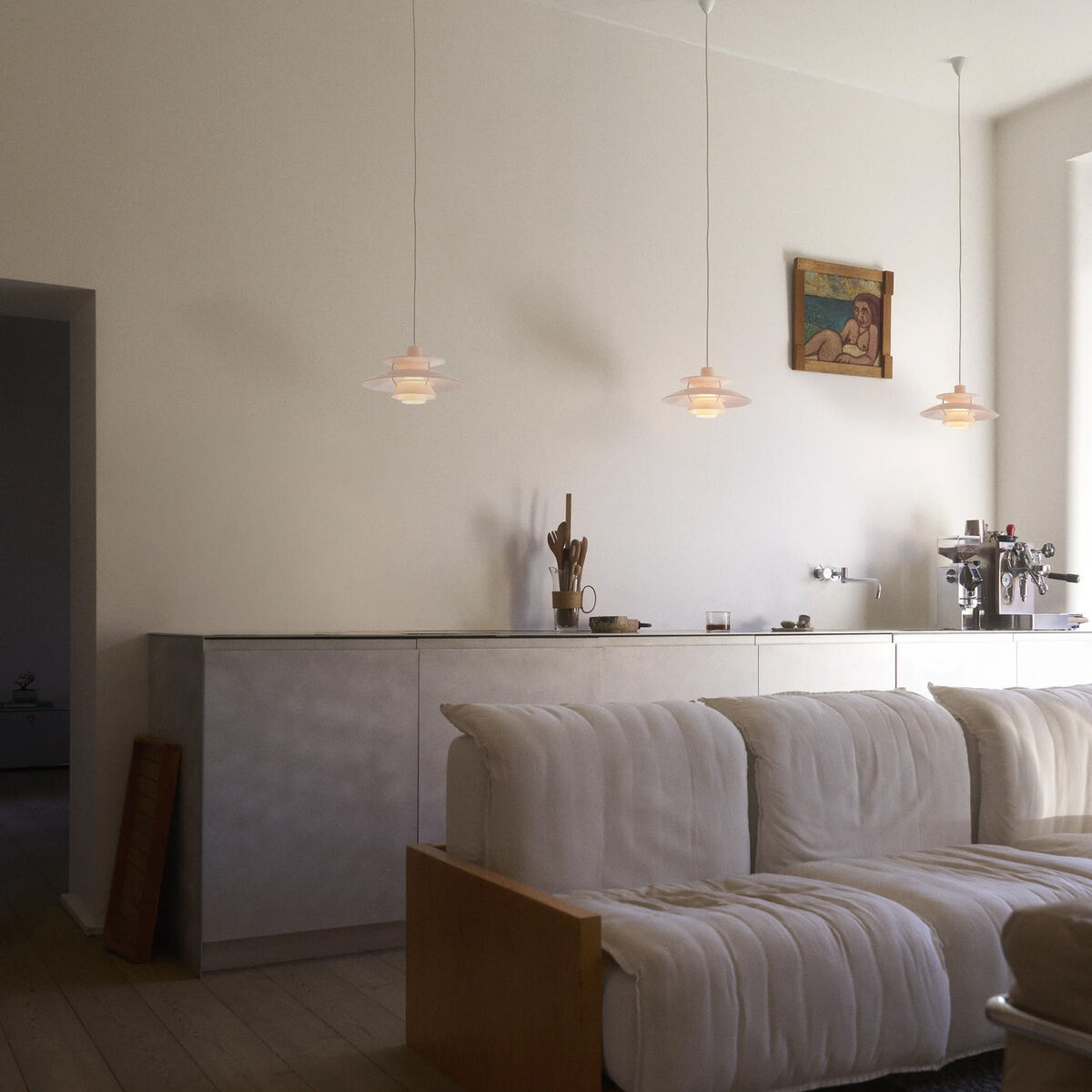 Moderne Wohnküche mit minimalistischem Design: Zwei Hängeleuchten »PH 5 320« von Louis Poulsen in Weiß und Pale Rose hängen über einer schlichten Küchenzeile, davor eine großzügige weiße Polstercouch. Helle Holzböden, viel Tageslicht und dezente Dekoration.