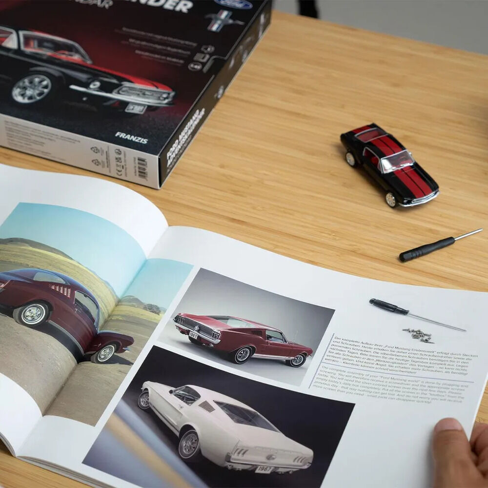 Auf hellem Holztisch liegen der teilweise sichtbare schwarze FRANZIS-Karton des Ford-Mustang-Adventskalenders, ein schwarz-rotes Mustang-Modellauto, ein kleiner Schraubendreher und ein geöffnetes Begleitbuch. Die Buchseiten zeigen Fotos roter und weißer Ford Mustang; eine Hand hält die rechte Seite.