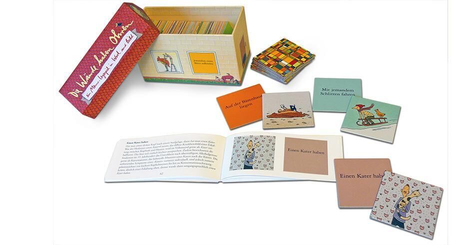Das Bild zeigt das Memo-Legespiel 'Wände haben Ohren', bestehend aus mehreren quadratischen Karten mit Illustrationen und Texten, sowie einem Buch. Die Karten und das Buch sind in einer rechteckigen Box mit rotem Deckel und bunten Seitenwänden verpackt.