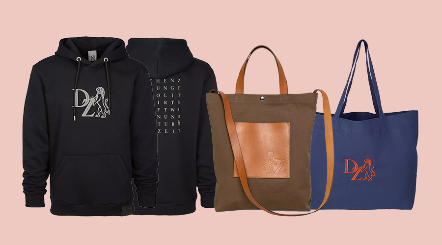 Schwarzer Hoodie mit weißem Löwen-Logo vorne und weißem Text auf der Rückseite, braune Stofftasche mit Lederapplikation und blaues Stofftragetasche mit orangefarbenem Löwen-Logo