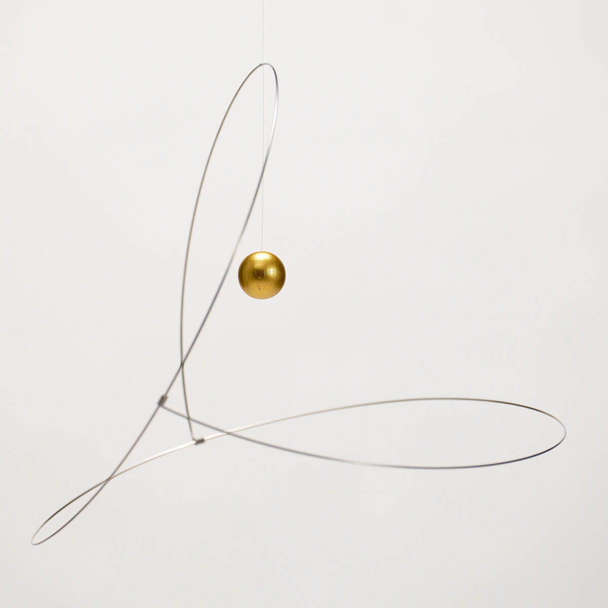 Das Mobile "Metamorphosis, Golden Planet" der Marke Flensted Hanging Art zeigt eine minimalistische Konstruktion aus feinem silbernem Draht, dessen Linien schwungvoll gekreuzt sind. Im Zentrum schwebt eine goldene Kugel. Der Hintergrund ist neutral weiß.