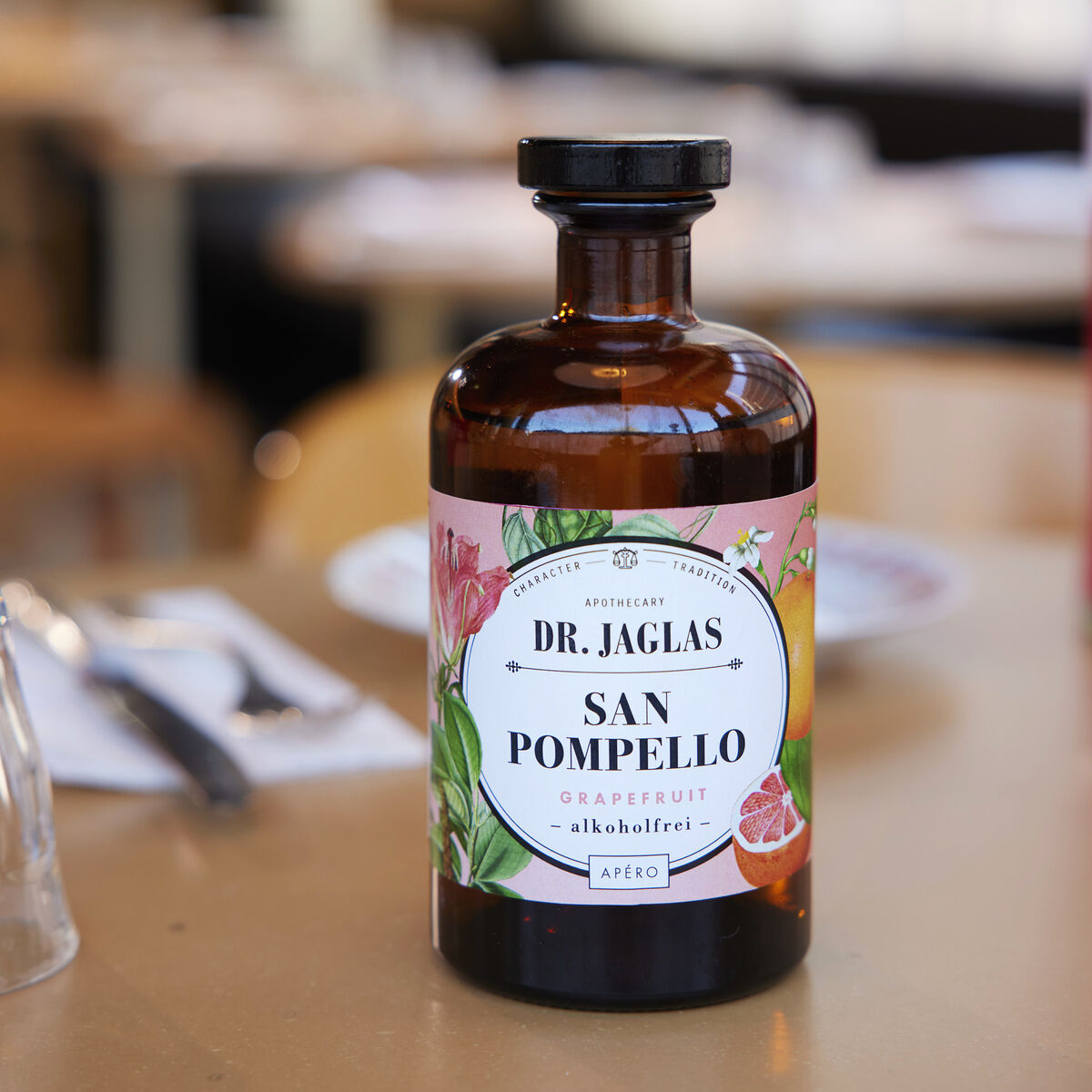 Eine Flasche San Pomello alkoholfreier Aperitif von Dr. Jaglas steht auf einem gedeckten Tisch mit unklaren Hintergrund. Das Etikett zeigt florale Muster und Grapefruit-Thematik.