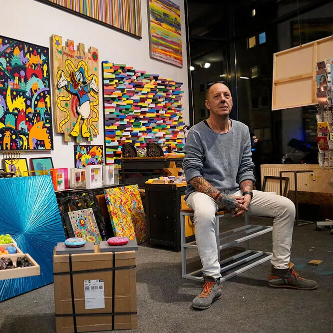 Der Kunstdruck »Love of my Life - Flowers« von Michael Pfannschmidt ist in einer kreativen Atelierumgebung zu sehen. Bunt gestaltete, teils abstrakte Werke hängen an weißen Wänden, darunter der limitierte, signierte Druck auf Aluminium, Maße 400 x 400 mm.