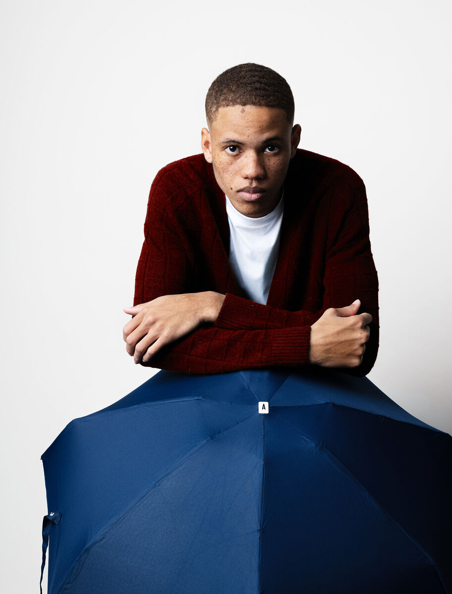 Ein Mann in roter Strickjacke und weißem T-Shirt hält einen navy-blauen Taschenschirm. Der Bildhintergrund ist weiß.