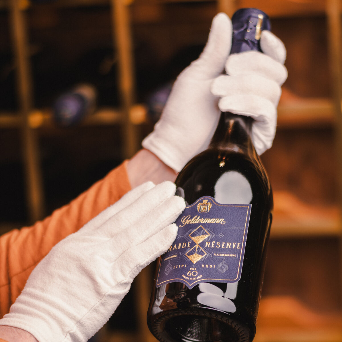 Eine stilvolle Präsentation einer Geldermann Grande Réserve Flasche, gehalten von Händen in weißen Handschuhen, vor einem hölzernen Weingestell. Das Etikett ist in edlem Blau und Gold gestaltet.