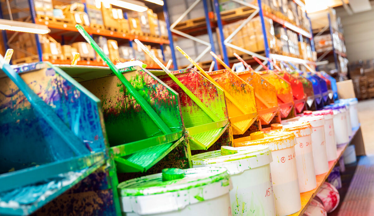 Eine Reihe von bunten Farbwannen sind in einem Lager aufgestellt, jede Farbe zur Hälfte gefüllt mit Farbresten. Es gibt Pinsel, die aus jeder Wanne herausragen. Die Farben umfassen Grün, Blau, Gelb, Rot und Orange. Im Vordergrund sind weiße Farbewannen zu sehen. Das Lager im Hintergrund zeigt Regale mit verschiedenen Artikeln.