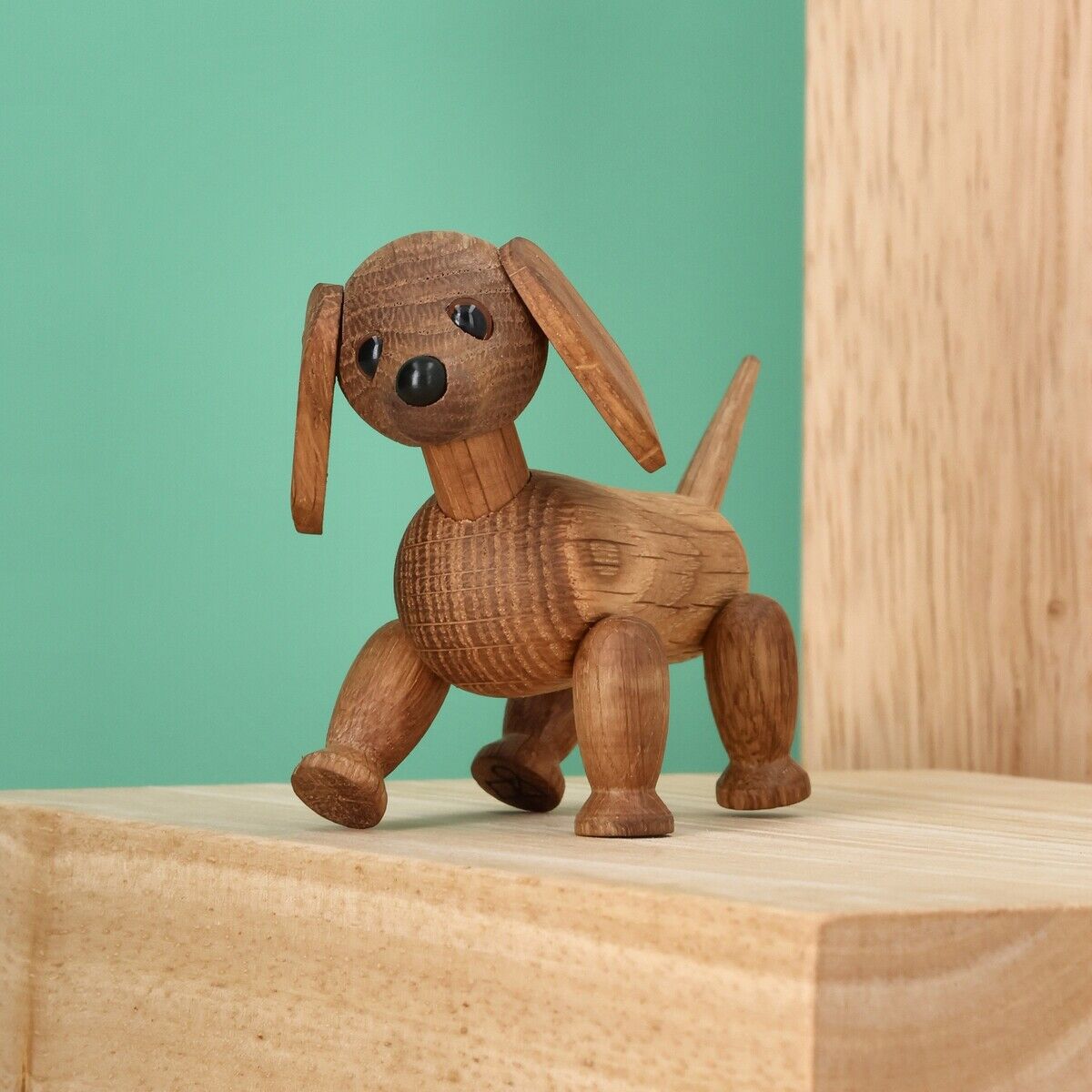 Die Holzfigur »Dackel Chico« von Spring Copenhagen steht auf einem hellen Holzpodest vor mintgrünem Hintergrund. Die detailreiche, handgefertigte Figur zeigt einen freundlichen Dackel mit beweglichen Ohren, Beinen und Schwanz. Modernes, minimalistisches Design.