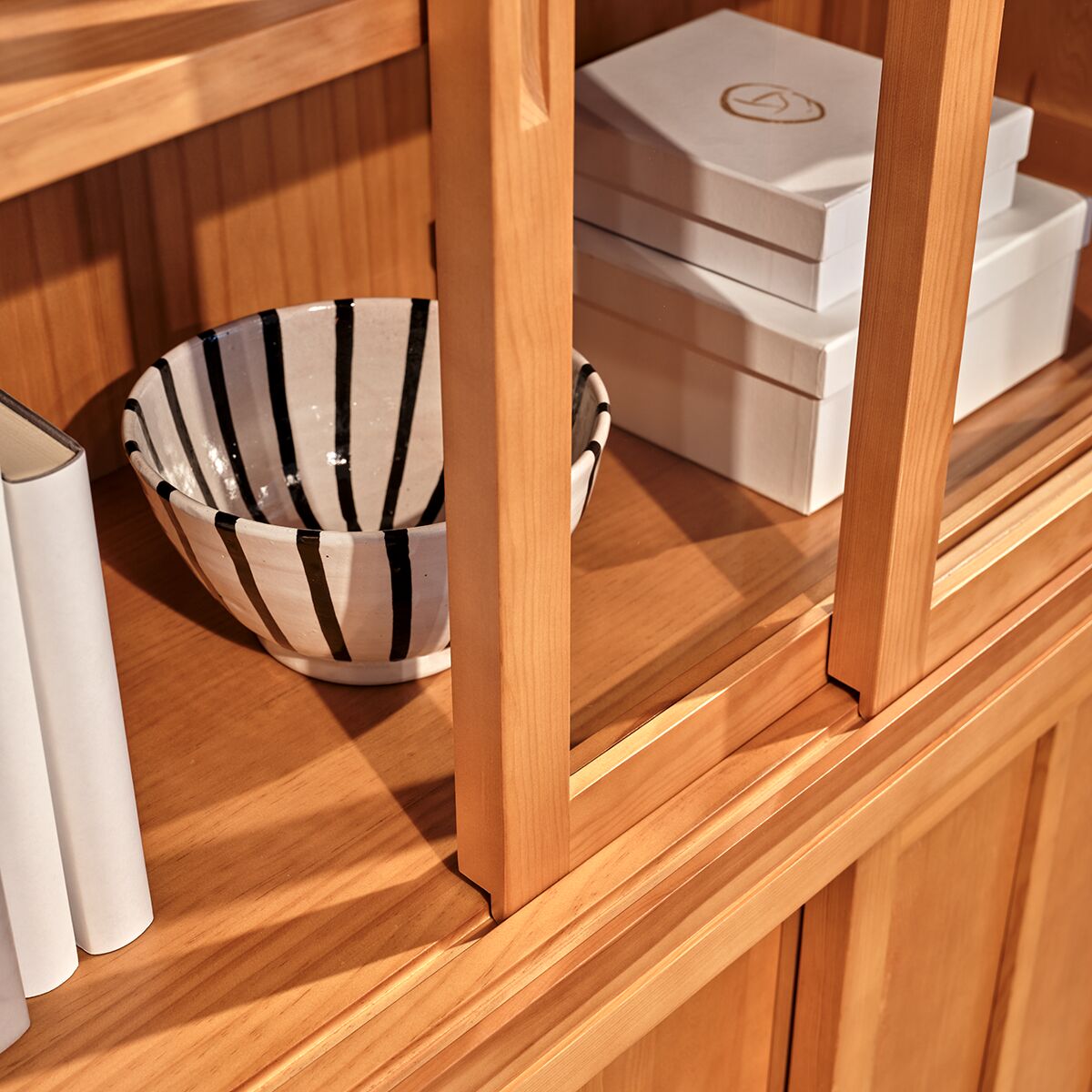 Detailansicht der honigfarben lasierten ZEIT-Sonderedition Vitrine »Multimodus« aus massivem spanischem Pinienholz mit klassischer Rahmenbauweise und Schiebetüren. Sichtbar sind gestapelte weiße Schachteln, einige Bücher und eine weiß-schwarz gestreifte Keramikschale im Inneren.