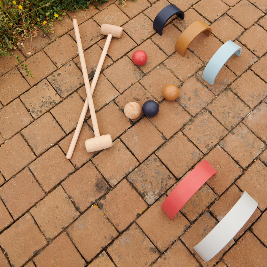 Ein Croquet-Set namens »Alen«, bestehend aus Holzkrokettschlägern, farbigen Bällen und gebogenen Toren, arrangiert auf einem Pflastersteinboden im Garten.