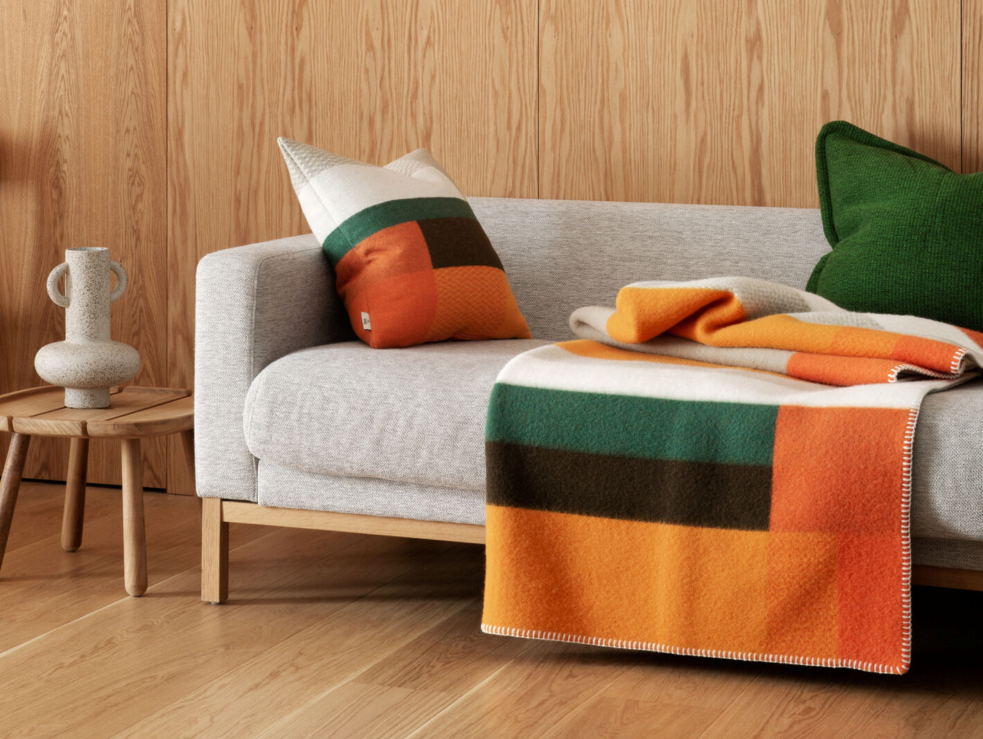 Hellgraues Sofa mit Kissen und Decke in Orange, Grün und Braun vor Holzpaneelwand, daneben kleiner Holztisch mit weißer Vase