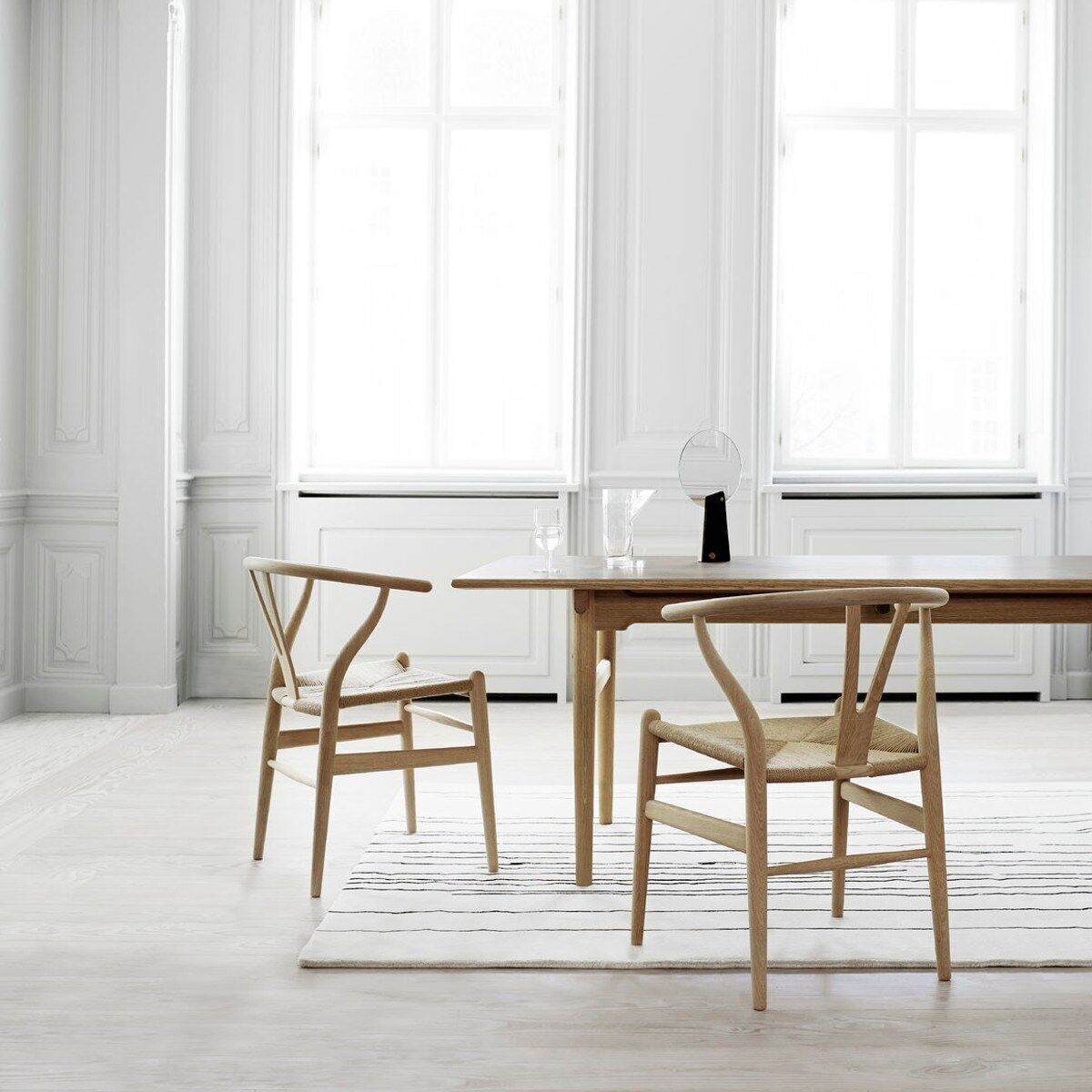 Der Wishbone Chair CH24 von Carl Hansen & Søn ist ein eleganter Holzstuhl mit einem minimalistischen Design. Er steht in einem hellen Raum mit großen Fenstern und einem weißen Teppich. Der Raum wirkt modern und einladend.
