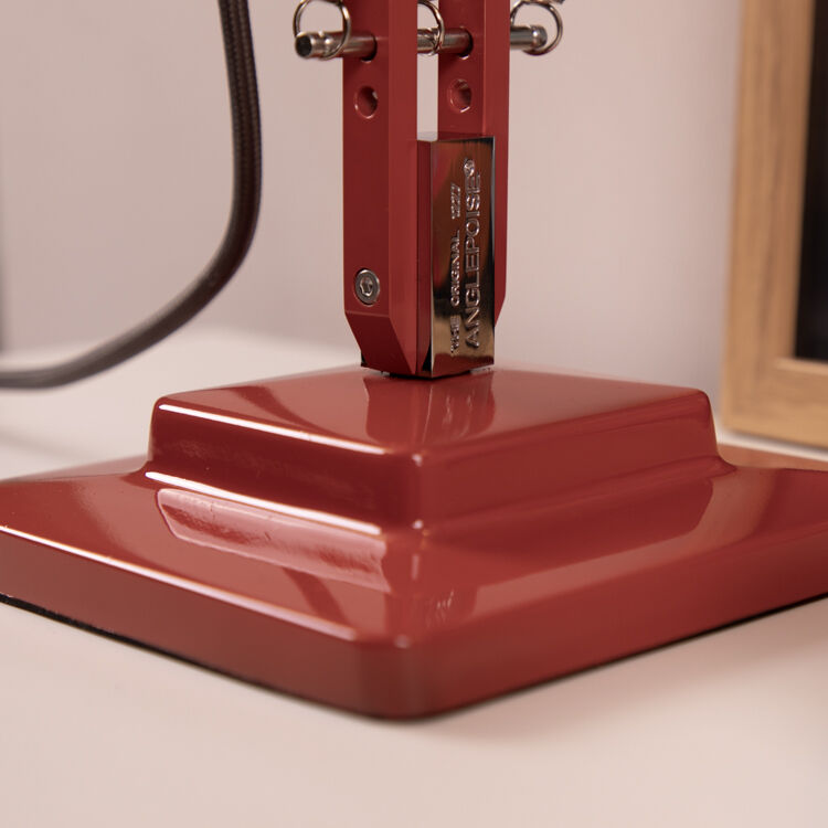 Nahaufnahme einer roten Tischleuchte vom Modell »Anglepoise Original 1227«, die Teil der ZEIT-Sonderedition ist. Die Lampe steht auf einer weißen Oberfläche neben einem Bilderrahmen und einer Schale.