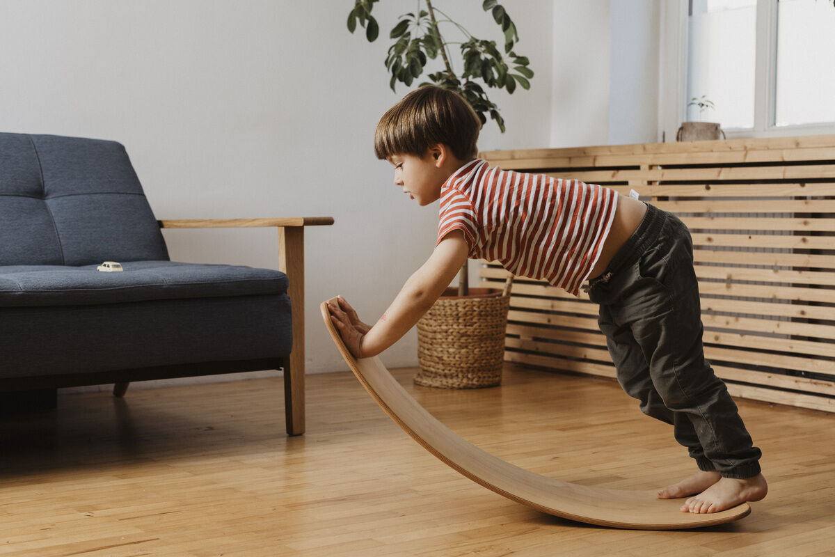 Ein kleines Kind spielt mit einem geölten Balance Board im Wohnzimmer. Der Boden ist aus Holz, und im Hintergrund sind ein blaues Sofa und eine Pflanze zu sehen.