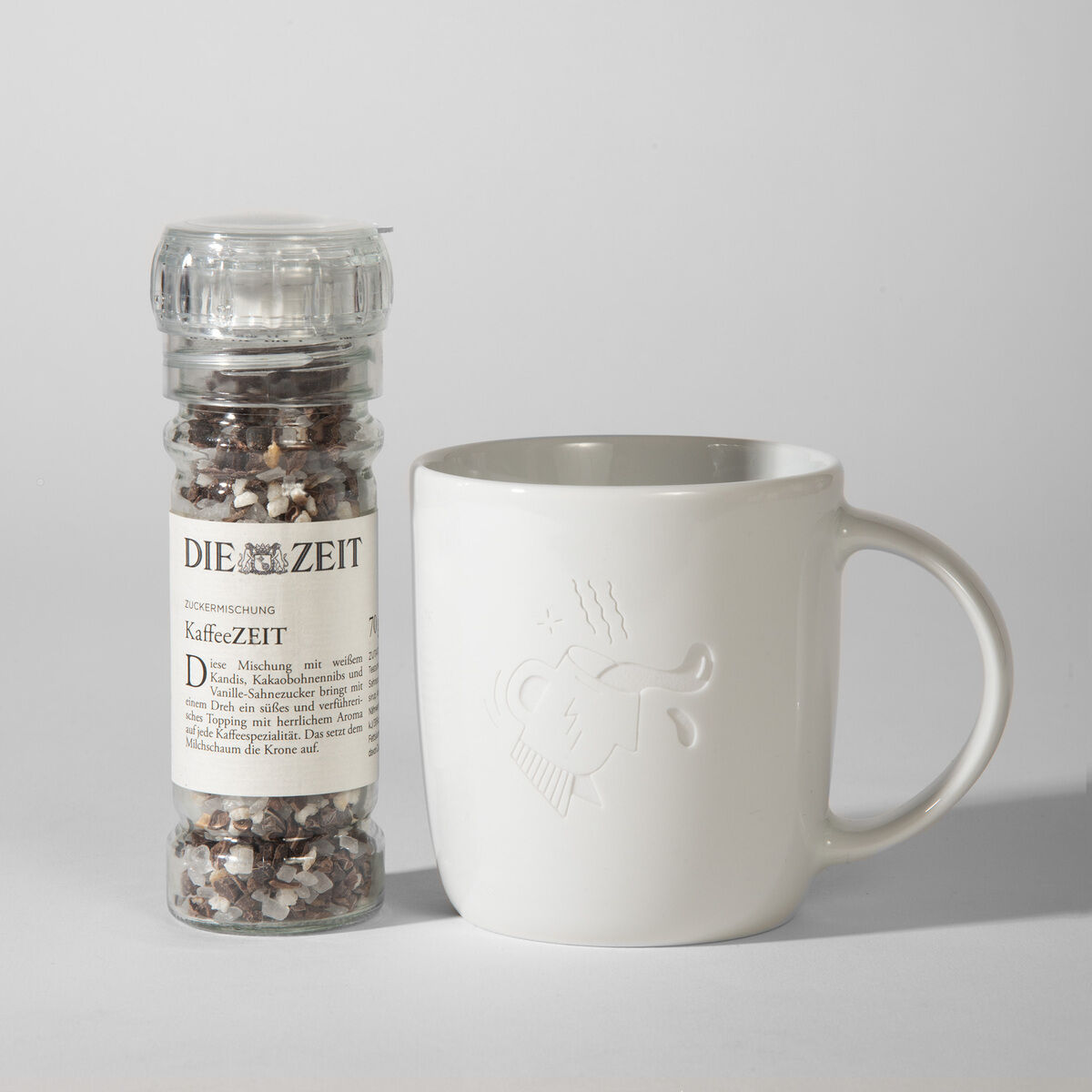 Das Bild zeigt ein Geschenkset, bestehend aus einer Kaffeetasse und einer Gewürzmischung namens KaffeeZEIT von der Marke Die Zeit. Die Tasse ist weiß und schlicht, mit einem dezenten Logo. Die Gewürzmischung ist in einem durchsichtigen Behälter und hat eine schwarze und weiße Farbgebung.