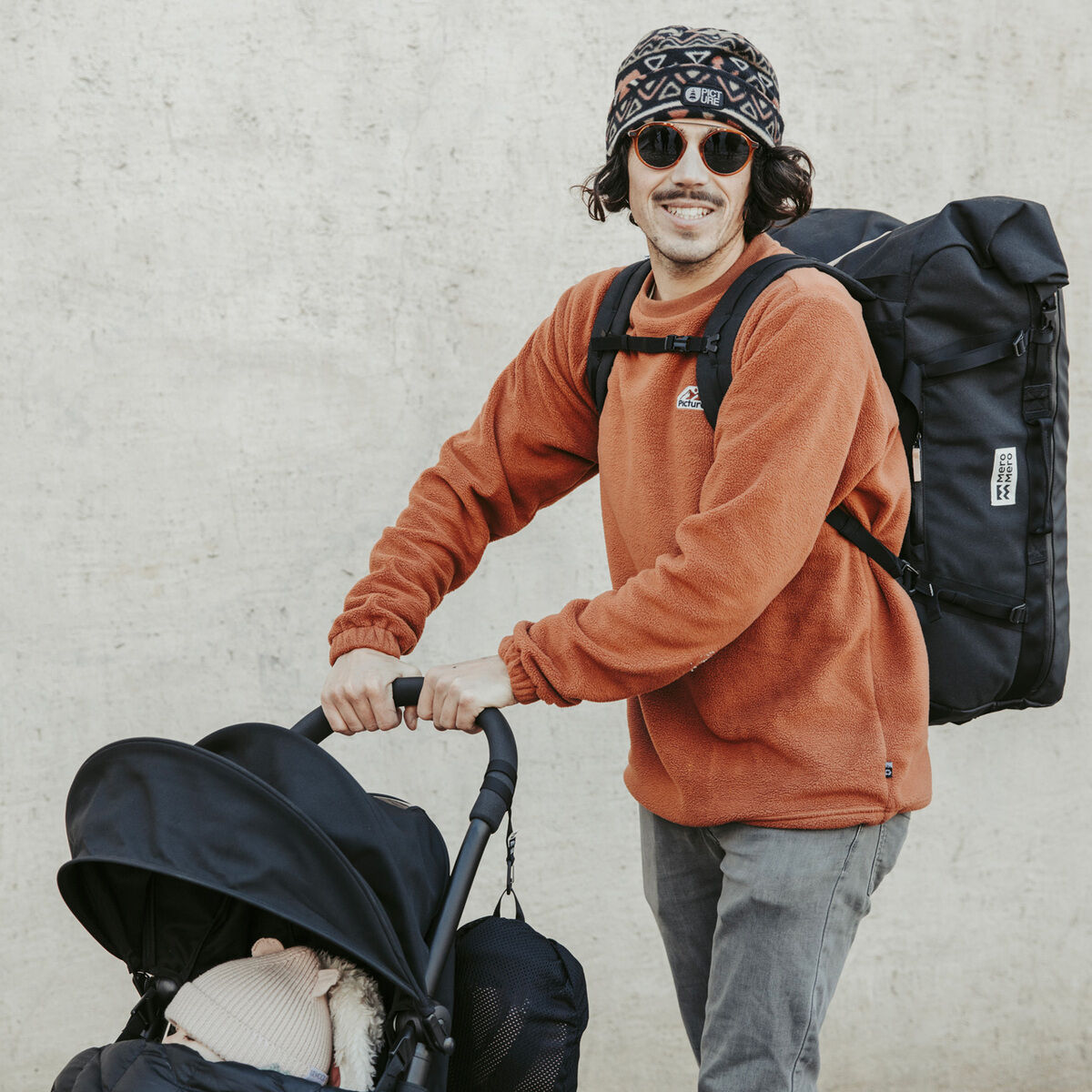 Ein Mann trägt eine schwarze Duffle-Tasche auf dem Rücken, während er einen Kinderwagen schiebt. Er trägt eine orangefarbene Jacke, eine gemusterte Mütze und Sonnenbrillen. Die Szene ist lässig und lässt auf eine Reise oder einen Familienausflug schließen.