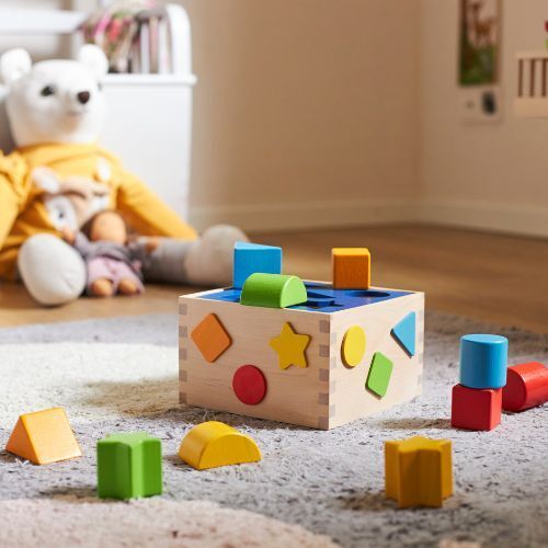 Ein buntes Holzspielzeug in Form einer Sortierbox befindet sich auf einem Teppich im Kinderzimmer. Verschiedene geometrische Formen, wie Quadrate, Dreiecke und Kreise, sind um die Box verteilt. Im Hintergrund sind eine weiße Plüschfigur und weitere Spielzeuge zu sehen.