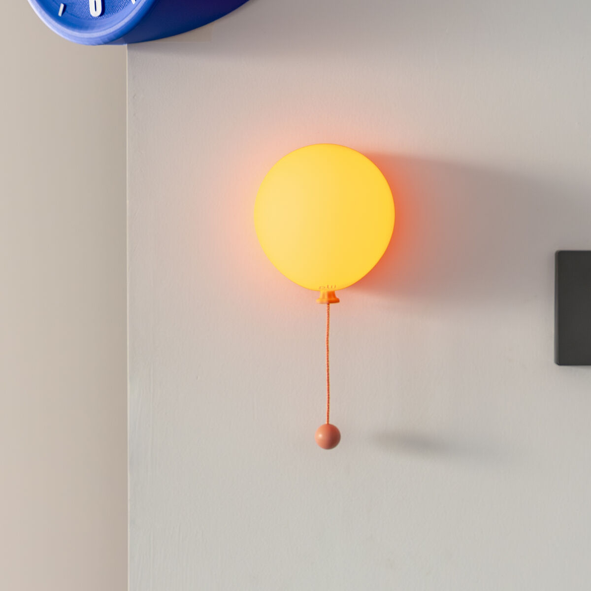 Auf einem hellen Hintergrund ist die Leuchte »Mimo Ballon« von Gingko Design Ltd in der Farbe honey glow als runde, gelb-orange leuchtende Ballon-Wandlampe mit Zugkordel zu sehen. Links oben ist eine blaue Wanduhr erkennbar.
