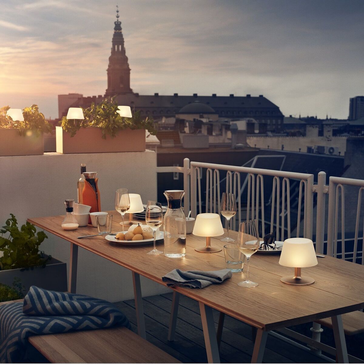 Solartischlampen SunLight auf einem Balkon mit Abendessen-Setting. Im Hintergrund eine städtische Skyline bei Sonnenuntergang, Pflanzen, Gläser und Geschirr auf einem Holztisch.