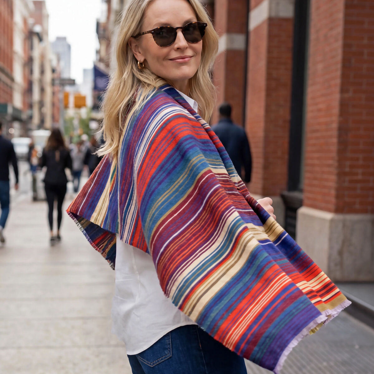 Eine Frau mit Sonnenbrille steht auf einem städtischen Gehweg vor rotbraunen Backsteingebäuden und blickt lächelnd zur Kamera. Über weißem Oberteil und dunkelblauer Jeans trägt sie den weit ausgebreiteten, weich fallenden Wollschal Ariana von Eagle Products mit unregelmäßigen Streifen in Rot, Blau, Violett, Grün und Gelb; im Hintergrund gehen Passanten.