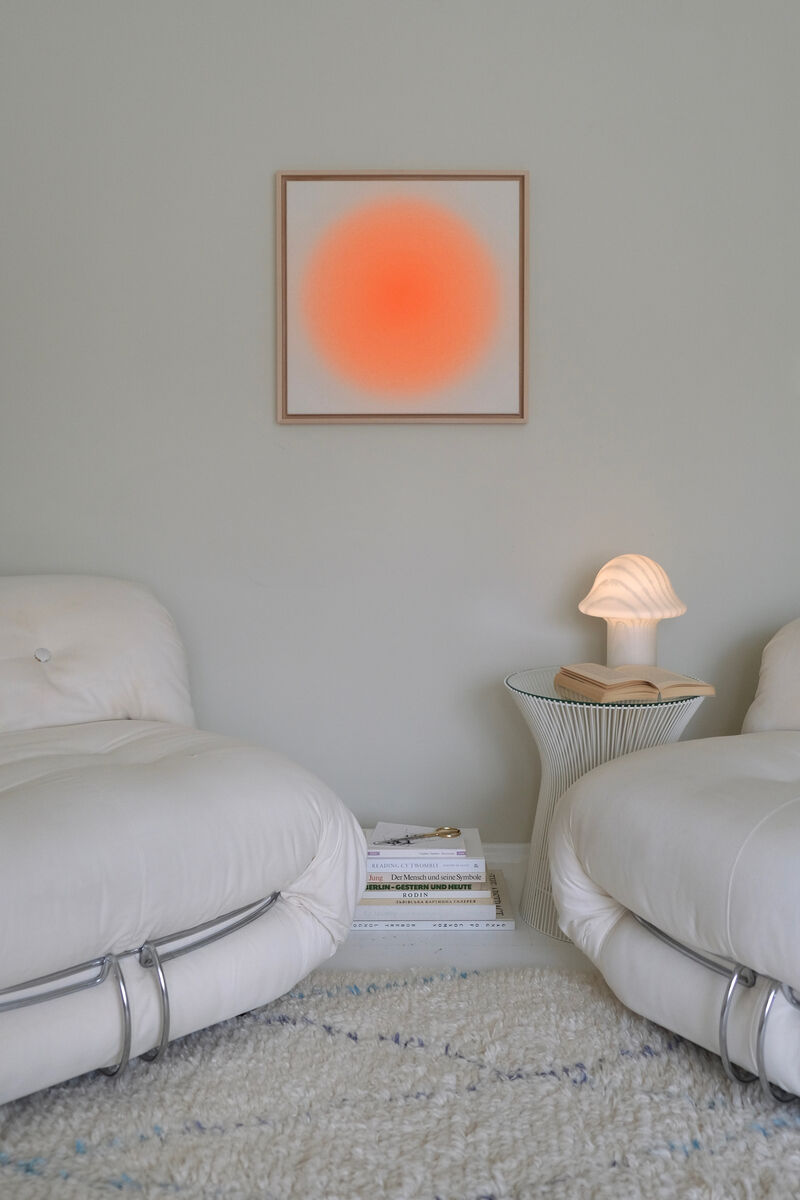 Zwei weiße Sessel auf einem hellen Teppich, zwischen ihnen ein kleiner Tisch mit gestapelten Büchern und einer Lampe. An der Wand hängt ein quadratisches Bild in einem schlichten Rahmen, das einen orangefarbenen Farbverlauf zeigt.